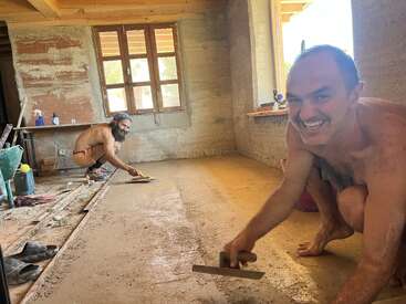 Two men are working shirtless, smiling while smoothing a freshly laid earthen floor inside a rustic, unfinished room with large windows and wooden ceiling beams.