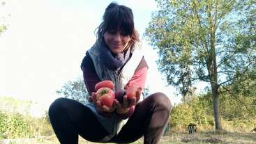 A woman squats outdoors, smiling and holding several fresh tomatoes and vegetables in her hands. She is surrounded by green trees and enjoys a sunny day.