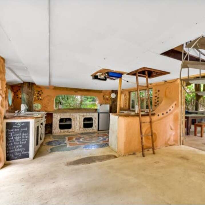This image shows a rustic, earthy kitchen with curved orange walls, mosaic details, large windows, open shelving, spiral staircase, and a cozy, artistic, handmade vibe.