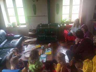 Una acogedora sala con niños y adultos reunidos en torno a una mesa de madera, disfrutando de las bebidas. La luz del sol entra por las ventanas; una estufa, libros y plantas completan el ambiente hogareño.