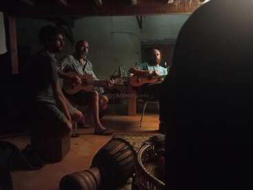 Tres hombres se reúnen en una habitación poco iluminada tocando guitarras acústicas e instrumentos de percusión, creando una atmósfera musical íntima y acogedora. Sombras e instrumentos llenan la escena.