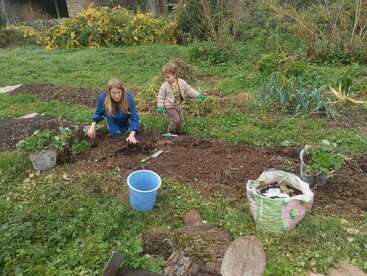 Una mujer y un niño trabajan juntos en un jardín, cuidando la tierra y las plantas con herramientas, rodeados de vegetación y cubos en un día nublado.