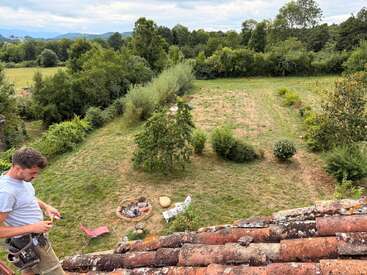 Un hombre está de pie en un tejado de tejas con vistas a un amplio patio trasero cubierto de hierba, con arbustos, árboles, una hoguera, sillas y rodeado de un denso bosque verde.