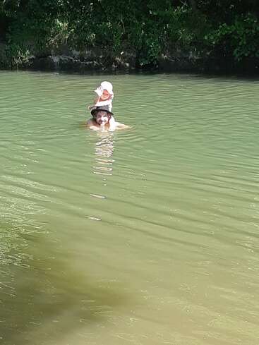 Um homem com um chapéu está nadando em um rio, carregando uma criança nos ombros. A criança está acenando, cercada por água verde e natureza.