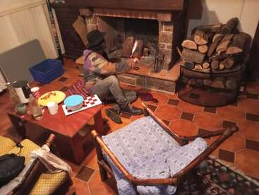 Uma pessoa com uma jaqueta colorida e chapéu preto coloca uma tora de madeira em uma lareira aconchegante. Sala rústica com pilha de lenha, lanches, bebidas e assentos confortáveis.