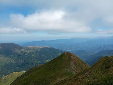 Esta imagem mostra uma deslumbrante paisagem montanhosa com colinas verdes, vales distantes e céu azul com nuvens dispersas. A vista é serena e ampla.
