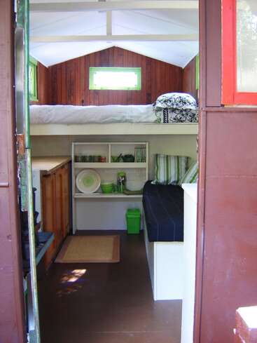 Esta casinha aconchegante tem uma cama elevada, paredes de madeira, uma cozinha compacta, prateleiras com louças, um banco com almofadas, tapetes e um interior luminoso e acolhedor.