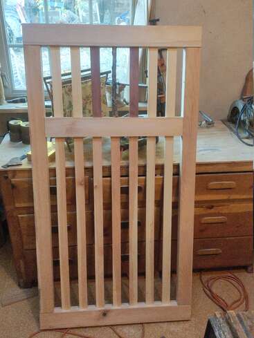 Un portail ou un panneau en bois se tient debout dans un atelier. Il se compose de lames verticales et d'une barre horizontale, entourées d'outils de travail du bois, de tiroirs et d'une fenêtre.