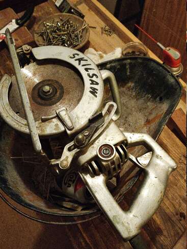 Une scie circulaire Skilsaw vintage repose dans un seau en métal, entourée de clous, d'outils et de fournitures sur un établi rustique en bois dans un atelier.