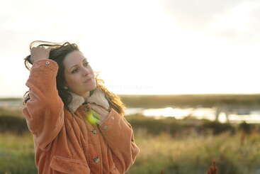 Eine Frau steht draußen auf einem Feld, hat eine warme orangefarbene Jacke an und berührt ihr Haar. Die Sonne scheint hell hinter ihr und schafft eine ruhige Atmosphäre.
