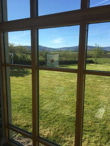 La imagen muestra una vista serena de un frondoso campo verde y montañas lejanas a través de una gran ventana con múltiples cristales, sobre un cielo azul despejado.