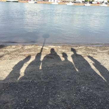 La imagen representa las sombras de cuatro personas de pie en una playa, con una masa de agua y un muelle al fondo, evocando una sensación de ocio y relajación.
