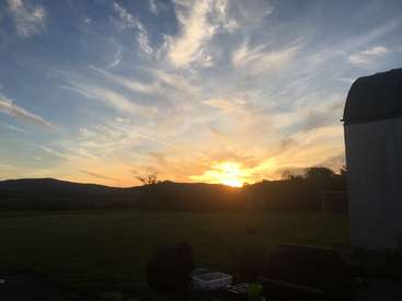 La imagen representa una serena puesta de sol con una caravana, montañas y un campo, sobre un cielo azul con nubes difusas.