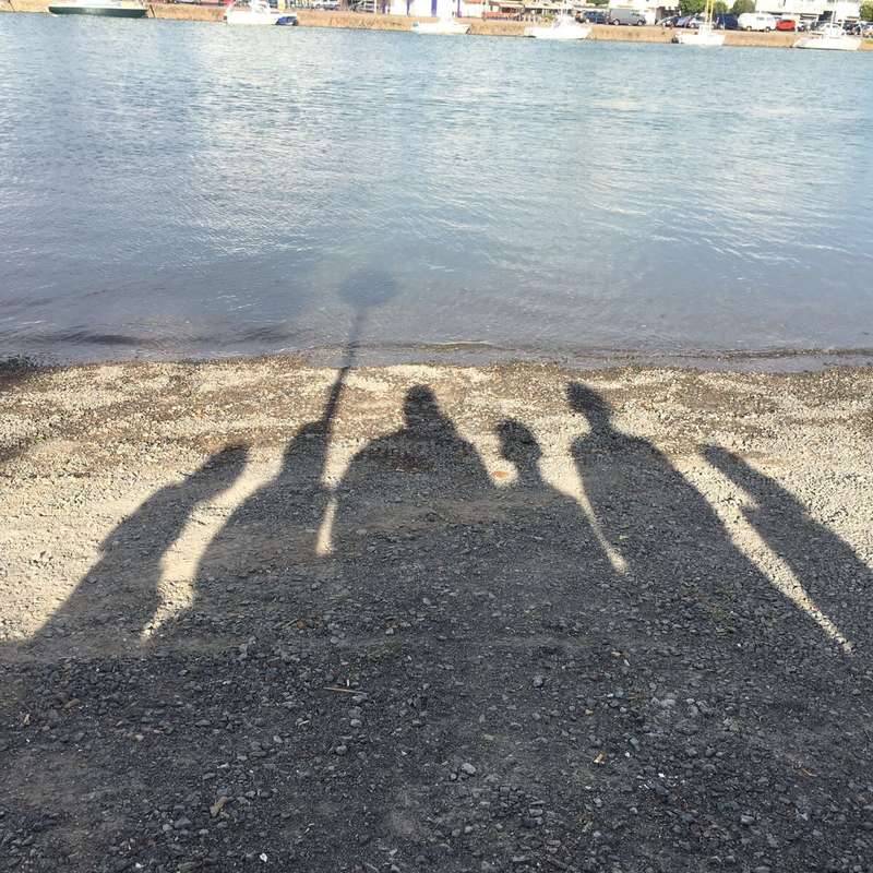 La imagen representa las sombras de cuatro personas de pie en una playa, con una masa de agua y un muelle al fondo, evocando una sensación de ocio y relajación.