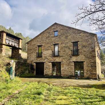 A rustic stone house with large windows and small balconies stands beside an old raised granary, surrounded by greenery, under a partly cloudy sky.