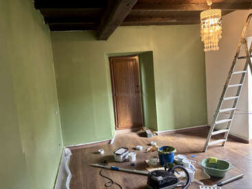 Ein Raum mit grün gestrichenen Wänden, einer Holztür und einer Holzdecke wird gerade renoviert. Malutensilien, Werkzeuge und eine Leiter liegen auf dem Boden verstreut. Oben hängt eine Lampe.