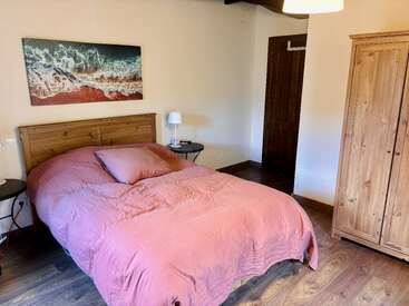 Ein gemütliches Schlafzimmer mit einem Doppelbett mit rosa Bettdecke, einem Kopfteil aus Holz, einem Kleiderschrank, Nachttischen, einer Lampe, einem Bild mit Meeresmotiv, Holzboden und neutralen Wänden.