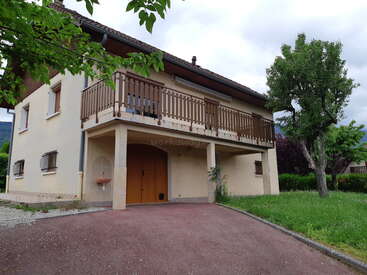The image depicts a two-story house with a balcony, featuring a driveway, trees, and a cloudy sky, set against a serene natural backdrop.