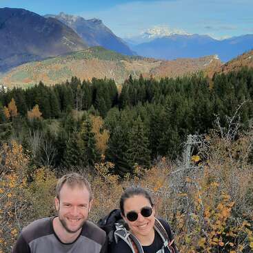 The image depicts a man and woman posing in front of a mountainous landscape, with trees and bushes in the foreground, set against a blue sky.