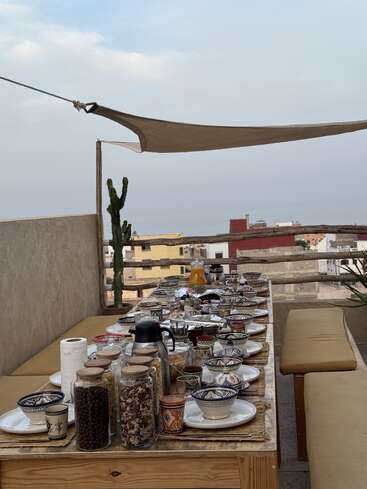 Uma bela mesa ao ar livre em um telhado, com cerâmica em estilo marroquino, potes de comida, bancos e sombreiros, com vista para uma cidade com prédios coloridos.