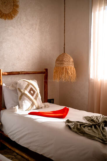 Um quarto aconchegante e iluminado pelo sol com uma cama de madeira, travesseiro geométrico, cobertor vermelho, luminária pendente de tecido e cortinas macias, criando uma atmosfera calorosa, convidativa e boêmia.