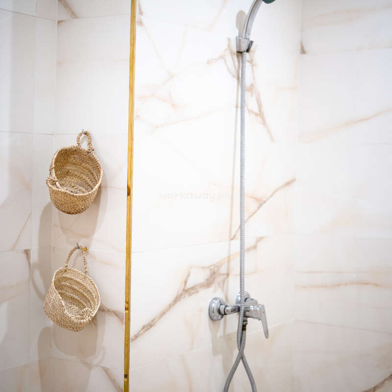 Esta imagem mostra uma área de chuveiro moderna com azulejos de mármore branco, um chuveiro de mão prateado e duas cestas suspensas de tecido presas à parede para armazenamento.