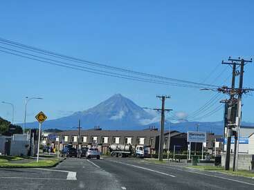 Une route dégagée traverse une zone suburbaine avec des voitures et des lignes électriques, tandis qu'une majestueuse montagne volcanique s'élève de manière spectaculaire en arrière-plan sous un ciel bleu.