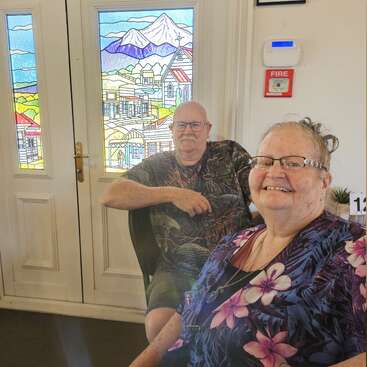 Un homme et une femme âgés sont assis à l'intérieur, souriants, devant de magnifiques portes vitrées représentant une ville colorée et des montagnes. Tous deux portent des lunettes et des chemises à motifs.