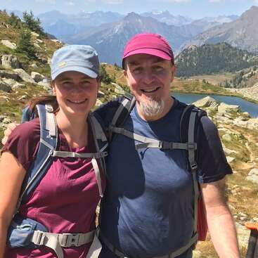 A smiling man and woman pose together while hiking in the mountains. Both wear backpacks and hats, with a lake and scenic peaks behind them.