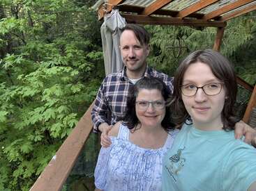 A family of three poses on a balcony surrounded by lush green trees. They are smiling, enjoying the outdoors, and appear happy and relaxed together.