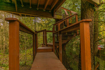 A wooden walkway with railings leads through a lush forest, connecting to a treehouse structure. Sunlight filters through green leaves, creating a peaceful, inviting atmosphere.