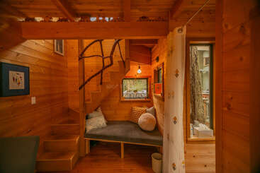 This cozy wooden cabin features warm lighting, a small staircase with a rustic branch handrail, a window nook with cushions, and charming, natural décor throughout.