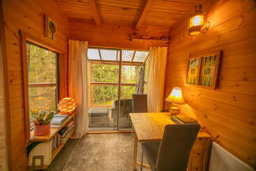 This cozy wooden room features a small desk with a lamp, two chairs, books, warm lighting, large windows, curtains, and a sliding door opening to greenery.