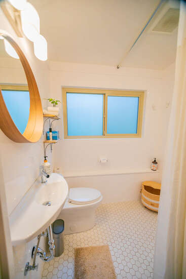 This is a compact, modern bathroom featuring a round mirror, white hexagonal tile floor, a toilet, small sink, window, wood bucket, and minimalistic decor.
