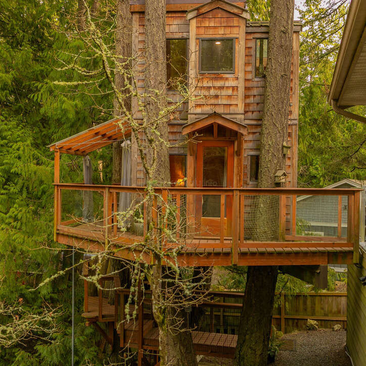 A charming wooden treehouse nestled among tall trees, featuring a wraparound deck, large windows, and rustic wooden shingles, blending harmoniously with the surrounding lush green forest.