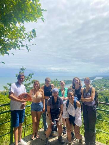 Um grupo de oito pessoas posa em um mirante panorâmico com vegetação exuberante, vista para o mar, céu nublado e alguns vestindo roupas de verão com câmeras e mochilas.