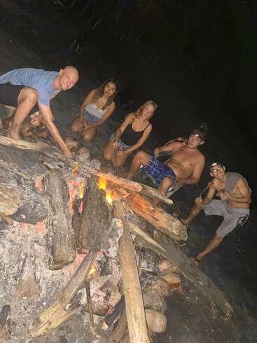 Cinco pessoas se reúnem ao redor de uma fogueira à noite, assando comida em palitos, sorrindo e curtindo a atmosfera ao ar livre, cercadas por troncos e brasas incandescentes.