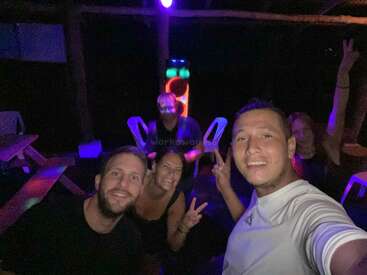 Cinco amigos posam para uma selfie à noite sob luzes coloridas, sorrindo e fazendo sinais de paz. Eles parecem estar curtindo uma festa ou reunião divertida.