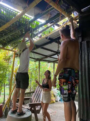 Três pessoas estão trabalhando juntas para consertar ou construir um telhado de bambu e folhas de palmeira. Duas delas estão em pé em superfícies elevadas, enquanto uma ajuda de baixo, ao ar livre.