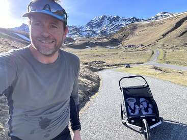 A imagem mostra um homem correndo em uma estrada de montanha, usando camisa cinza e boné, com um carrinho de bebê ao seu lado, em um pitoresco cenário montanhoso.
