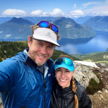 The image depicts a man and woman posing for a selfie on a mountain overlooking a lake, both wearing hats and jackets, with a serene landscape in the background.