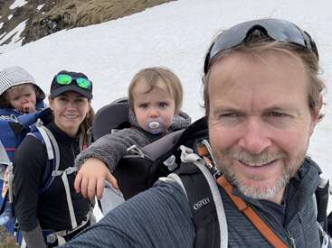 Uma família de quatro pessoas faz caminhadas em montanhas nevadas. Os pais carregam seus filhos pequenos em mochilas. Todos estão agasalhados, sorrindo e aproveitando a aventura ao ar livre.