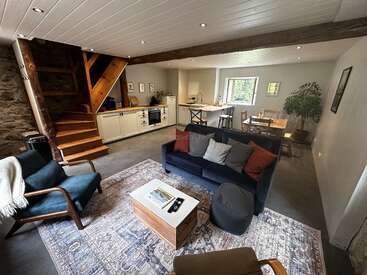 Cet espace de vie ouvert et confortable comprend une cuisine moderne, une table à manger, des sièges confortables, un escalier en bois, une plante d'intérieur, une lumière naturelle et des poutres apparentes rustiques.