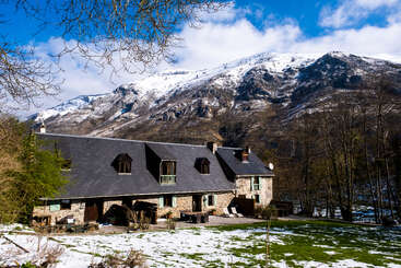 Une maison rustique en pierre repose paisiblement au pied de montagnes enneigées, entourée d'arbres, de plaques de neige, d'herbe verte et sous un ciel d'un bleu éclatant.
