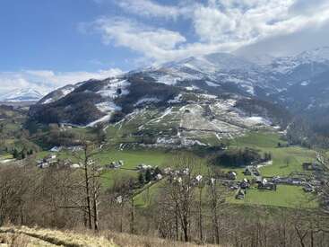 Un paysage de montagne pittoresque présente des pentes enneigées, des champs verts, des arbres épars et un petit village niché en contrebas, sous un ciel bleu partiellement nuageux. Un paysage paisible.