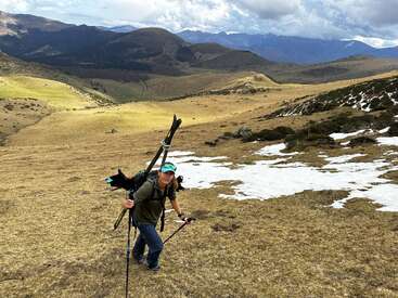 Une personne monte une pente de montagne herbeuse en portant des skis et en utilisant des bâtons de randonnée, avec des plaques de neige et des collines spectaculaires à l'arrière-plan.