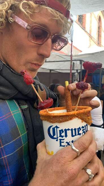 A man with curly hair, sunglasses, and scarf enjoys a beer in a cup decorated with chili powder, tamarind candy, and skewered fruit, at an outdoor market.