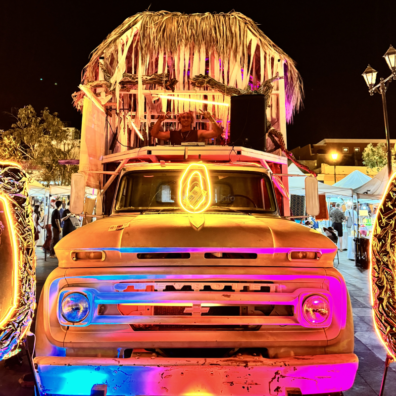 An old Chevrolet truck decorated with colorful neon lights and a thatched roof, hosting a DJ on top, is parked at a lively nighttime street market.