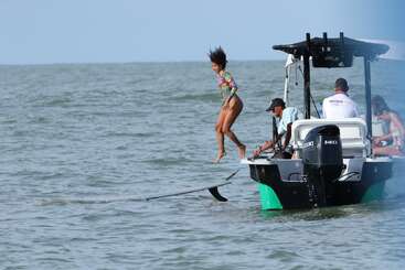 A woman jumps off a boat onto a hydrofoil board in the ocean, while three people on the boat watch her. The weather is clear and sunny.