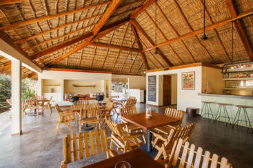 This open-air restaurant features wooden tables and chairs, a thatched roof, a bar area, tropical decor, natural sunlight, and relaxed, inviting atmosphere, perfect for dining.